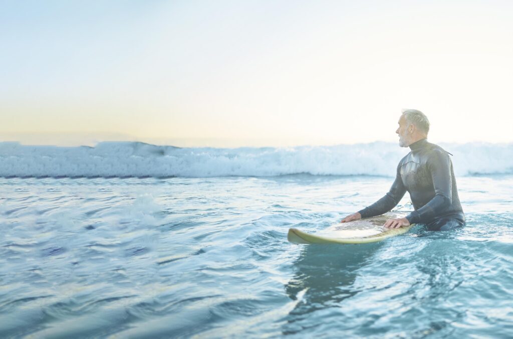 AMRA Swimming, beach and old man in water surfing enjoying a summer holiday vacation his retirement in Los Angeles, USA. Fitness, healthy and senior person waiting for ocean waves on his surfboard at sea